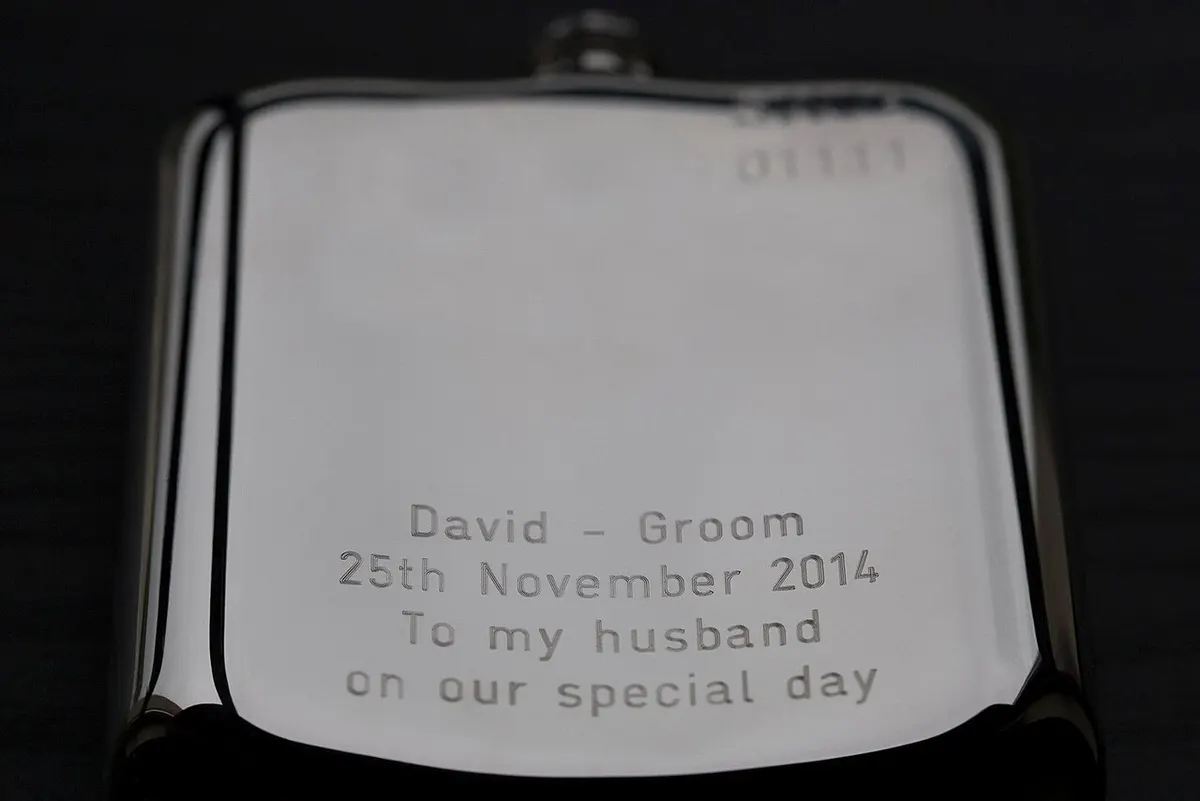 A stainless steel hip flask with an engraved lid on a wooden bar top